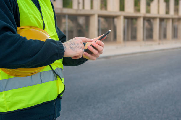 istock Close-up of a laborer looking at a phone while holding a yellow hard hat with a construction site in the background 1461688539