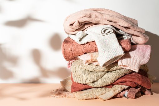 Messy pile of textured woolen sweaters in pastel beige, pink and orange colors against sunlit white background. Autumn and winter fashion aesthetic.