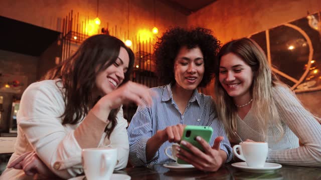 Group of young women having fun sharing media with an cellphone. Three girls looking to the smartphone on a coffee shop, restaurant or bar. Female students buying on a market place using a mobile phone with internet indoors.