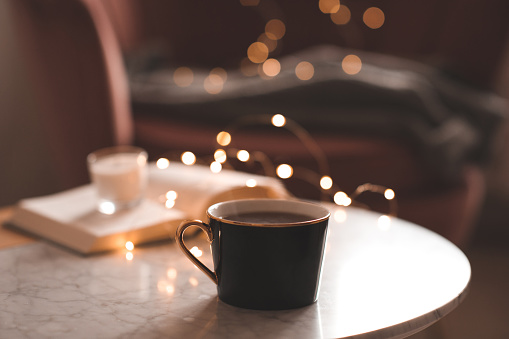 Cup of coffee with stack of paper books and scented candle over glowing lights in bedroom with armchair and knit clothes indoor. Cozy home atmosphere.