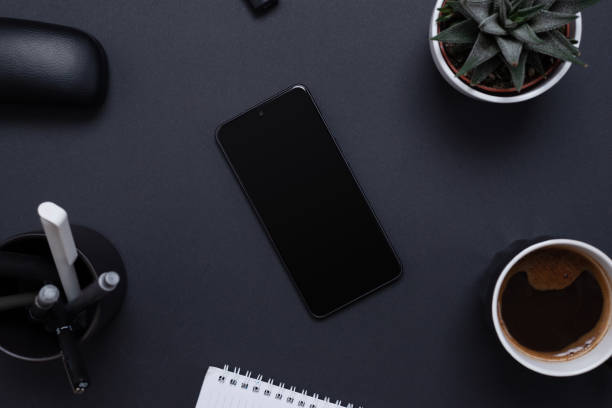 istock Minimalist and organized work desk with a blank smart phone surrounded by a plant, pens, coffee and notepad. Blank screen for mockup app presentation. Office work desk composition, top view, flat lay 1461784940