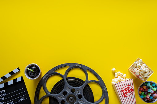 This is a conceptual photo relating to going out to the movies or watching a movie at home. There are two old retro movie reels on a bright yellow background with two bags of popcorn and hand made cinema tickets. There is a lot of space above for copy.