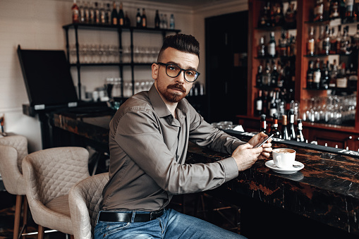 Middle-aged man in formal wear and glasses sitting in the bar and drinking his morning coffee.