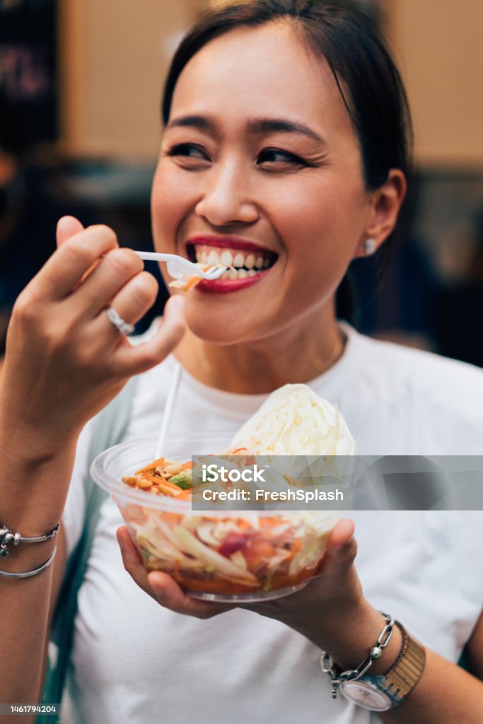 A Happy Beautiful Woman Holding Eating Some Healthy Salad A smiling Asian female looking away while enjoying street food. Bowl Stock Photo