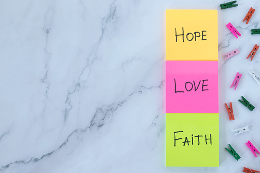 Love, hope, and faith handwritten words on colorful notes with decorative clothespins on white marble background. Top table view. Copy space. Christian biblical concept, 1 Corinthians 13 bible book.