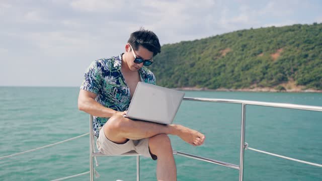 Asian young handsome businessman use laptop computer working in yacht. Attractive male freelance tourist hang out and typing work project on notebook while catamaran boat sailing during summer sunset.