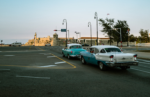 Havana, Cuba - December 12th 2022: Classic vintage cars driving along the Malecon at old Havana during dusk hour. Sunset lighting the face of a cuban girl in the car. Malecon is iconic spot for tourists, also the most popular place for cubans to hang out.