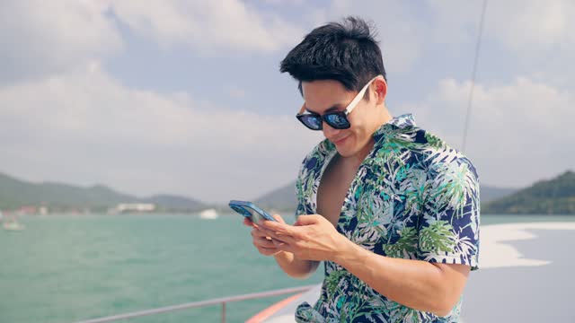 Asian young attractive man tourist use mobile phone chatting in yacht. Attractive male traveler online messenger on smartphone with friend while catamaran boat sailing during summer sunset yachting.