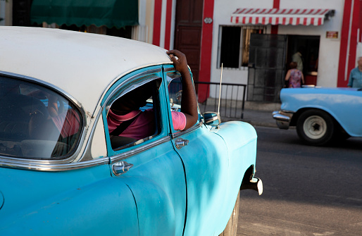 An unrecognizable old car driving on a street in Havana, with the passenger's arm visible