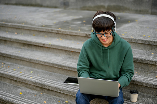 Smart and happy young Asian male college student wearing wireless headphones and glasses using laptop computer on campus building outdoor stairs, remote working school project.