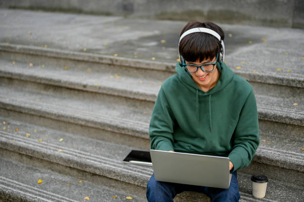 estudante universitário asiático inteligente usando fones de ouvido, usando laptop nas escadas ao ar livre do campus - male education college student nerd - fotografias e filmes do acervo