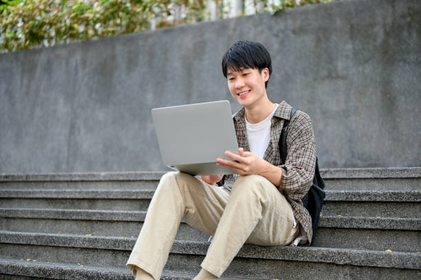 istock Happy Asian male college student working on his school project, using laptop on campus outside stairs. 1461922928