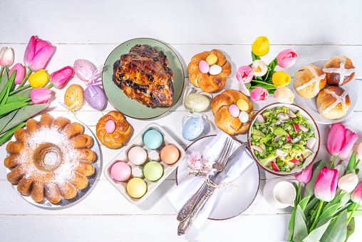 Festive served Easter dinner or brunch table, with Easter egg, tulip flower bouquet, traditional Easter foods - cake, cross buns, wreath bread, glazed ham and spring salad, white table background