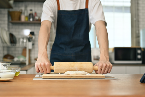 Man wearing aprons preparing homemade pastry, kneading dough with a rolling pin on wooden table in kitchen.