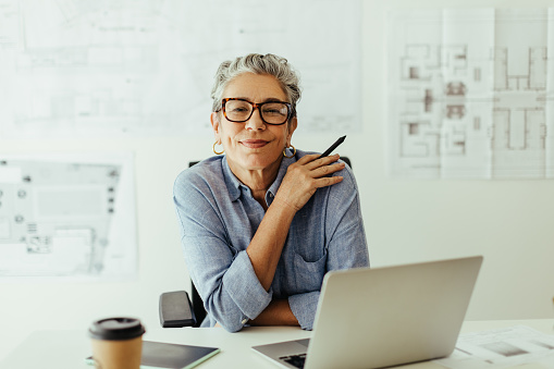 Mature female designer looking at the camera in her office, confident in her expertise and style of creativity. Senior design professional using a laptop and a graphics tablet to put her skills to work.