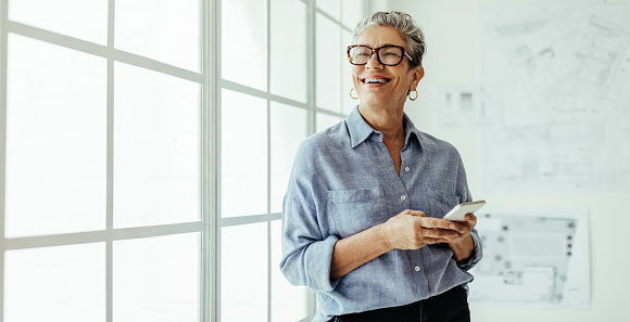 Mature business woman stands in an office, holding a smartphone and looking outside the window with a smile. Female designer using mobile technology to stay connected with her colleagues and clients.