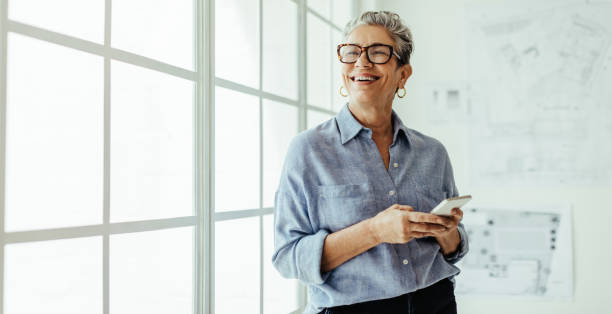 Mature business woman smiling and using a mobile phone in her office Mature business woman stands in an office, holding a smartphone and looking outside the window with a smile. Female designer using mobile technology to stay connected with her colleagues and clients. happy using phone stock pictures, royalty-free photos & images