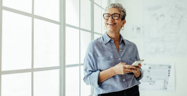 mature business woman smiling and using a mobile phone in her office - olhar para longe imagens e fotografias de stock