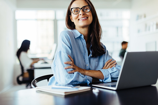 Female web developer smiling at the camera while sitting with a laptop in an office. Mature business woman working in a creative office.