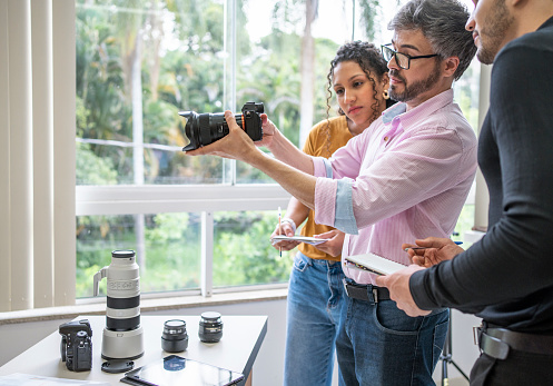 Professional photographer teaching two young assistants taking notes about camera equipment during a class in his studio