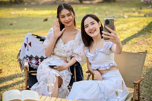 Two beautiful young Asian women in lovely dresses enjoying picnic in the green park together, taking selfies with their phone. besties, best friend, relationship