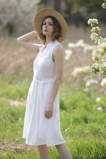 Spring fashion campaign. Outdoor portrait of female model in white linen dress and summer straw hat enjoying in nature during beautiful summer day