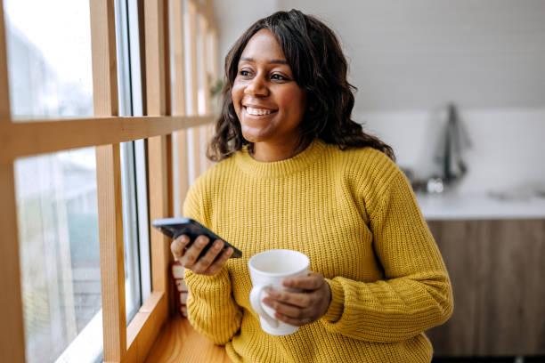 istock Beautiful woman using phone while having coffee while looking through the window in apartment 1462069889