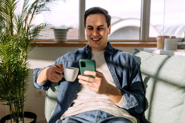istock Young smiling casually clothed man having coffee while text messaging and sitting on the sofa at his apartment 1462070204
