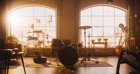 Establishing Shot: Music Rehearsal Studio in Loft Room with Drum Set in the Middle. Stylish Interior with Two Big Windows, Cozy Sofa, Shelves and Plants. Sunny Bright Day and Urban View.