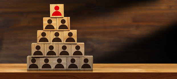 Leadership or business motivation concept. Group of brown people icons and one red on the top, on wooden blocks forming a pyramid, above a wooden table or desk with copy space.