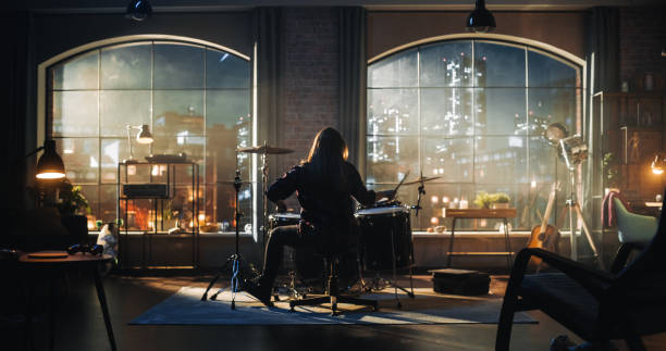 istock Person Sitting with Their Back to Camera, Playing Drums During Rehearsal in a Loft Studio with Sunlight. Drummer Practising Alone Before a Live Concert on Stage. Evening or Night Session. 1462111503