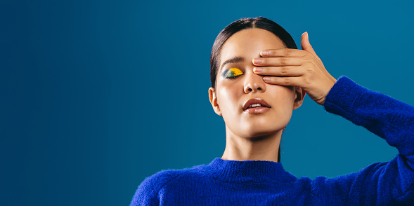 Woman in a blue sweater covers her eye to highlight her striking graphic eyeliner. Glamorous young woman wearing a bold eye makeup look in a studio.