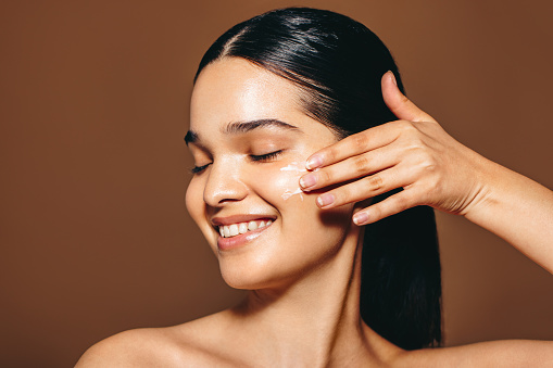 Young woman with perfect skin applies facial cream, she smiles as she makes use of this beauty product with is an essential part of her daily skincare routine. Woman in her 20’s caring for her skin.