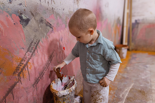 An adorable toddler boy with blond hair working in his father's workshop. He is learning how to paint a large canvas.