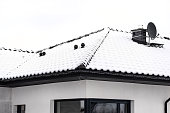 The roof of a single-family house is covered with snow against a cloudy sky, visible ceramic ventilation fireplace and a satellite dish.