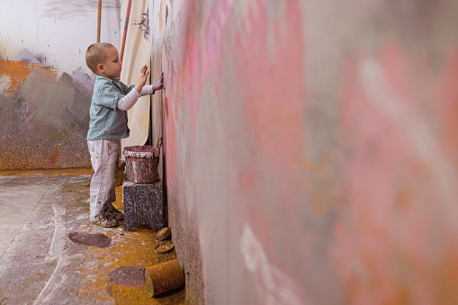An adorable toddler boy with blond hair working in his father's workshop. He is learning to paint the wall.
