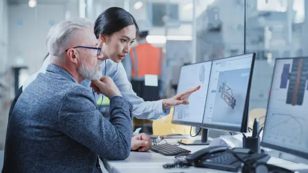 Photo of Factory Office Facility: Industrial Engineers Drafting Blueprints of a Heavy Industry Machine Parts on a Computer CAD Software. Asian Female Technician Discusses Work with Middle Aged Designer.