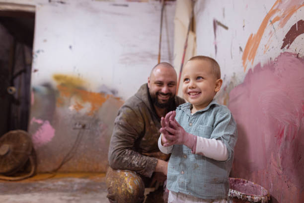 A shot of an adorable happy toddler boy with blue eyes, having fun with his dad in a workshop. They are painting a wall in pink color.