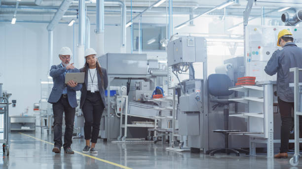 istock Two Diverse Heavy Industry Engineers in Hard Hats Walking with Laptop Computer and Talking in a Factory. Footage of Two Manufacturing Employees at Work. 1462137939