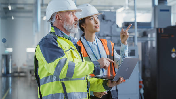 istock Two Diverse Professional Heavy Industry Engineers Wearing Safety Uniform and Hard Hats, Working on Laptop Computer. Asian Technician and Worker Talking Next to a Factory Machine. 1462138538