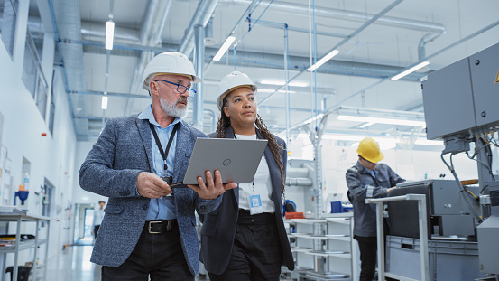 Dos ingenieros profesionales de la industria pesada con cascos en la fábrica. Caminando y discutiendo la instalación de la máquina industrial, trabajando en la computadora portátil. Gerente y Técnico Afroamericano en el Trabajo. photo