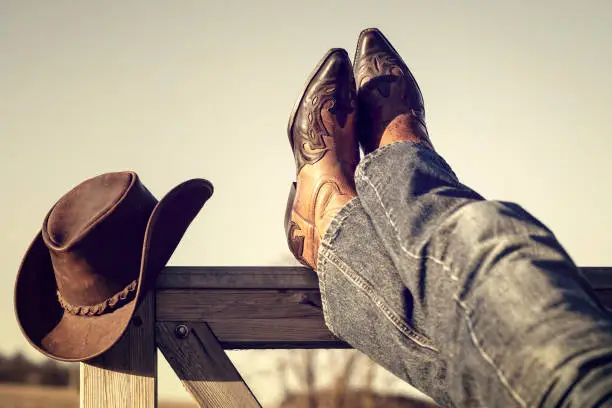 Cowboy boots and hat with feet up on stables gate at ranch resting with legs crossed, country music festival live concert or line dancing concept