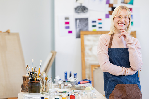 Young woman is standing behind working table in her painting studio.  She is posing for the camera.