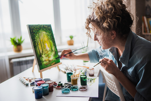Beautiful woman artist sits in front of a painting and paints with brushes on canvas at home