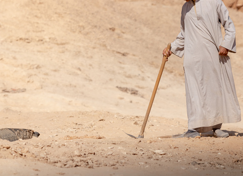 Unrecognizable hard working arab man with djellaba using shovel for digging in the sand sorrounded by desert and dunes