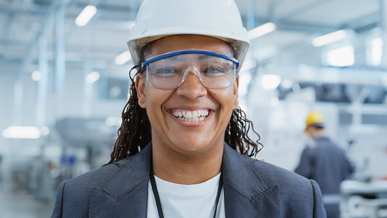 Close Up Portrait of a Happy and Smiling Middle Aged African Female Engineer in White Hard Hat Standing at Electronics Manufacturing Factory. Black Heavy Industry Specialist Posing for Camera.