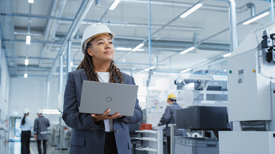 Portrait of a Black Female Engineer in Hard Hat Standing and Using Laptop Computer at Electronic Manufacturing Factory. Technician Thinking About Daily Tasks and Working on Project Pipeline.