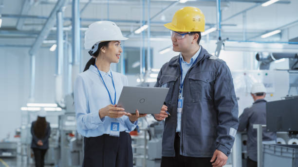 istock Two Diverse Young Heavy Industry Engineers in Hard Hats Walking with Laptop Computer and Talking in a Factory. Two Manufacturing Employees at Work in Research and Development Facility. 1462148693