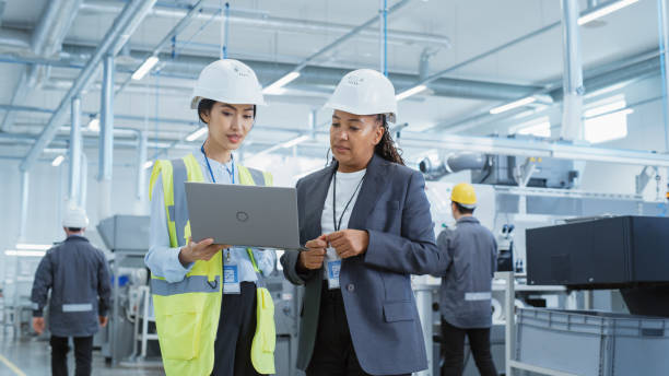 Two Female Heavy Industry Employees in Hard Hats at Factory. Discussing Job Assignments at Industrial Machine Facility, Using Laptop Computer. Asian Engineer and African American Technician at Work. dos empleadas de la industria pesada con cascos en la fábrica. discutir las asignaciones de trabajo en las instalaciones de máquinas industriales, utilizando computadoras portátiles. ingeniero asiático y técnico afroamericano en el trabajo.