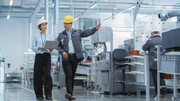 istock Two Young Asian Heavy Industry Engineers in Hard Hats Walking with Laptop Computer and Talking in a Factory. Two Manufacturing Employees at Work in Production Facility. 1462149085